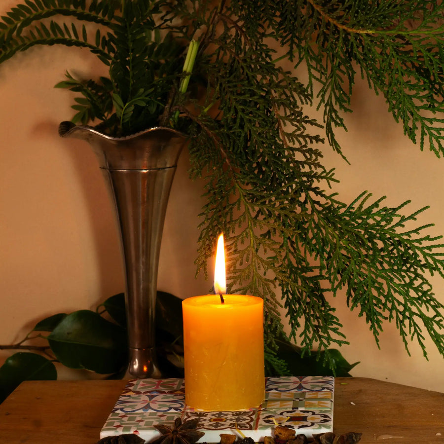 A tall, slender metallic vase with a flared rim holds green foliage beside a lit orange candle.